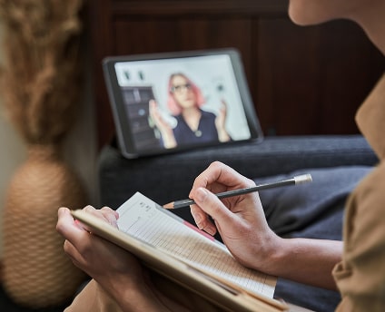 Person taking notes on a clipboard while attending a virtual therapy session on a tablet.