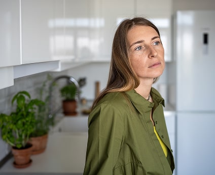 Woman in a green shirt standing in a kitchen, looking thoughtful and concerned, representing stress or anxiety.